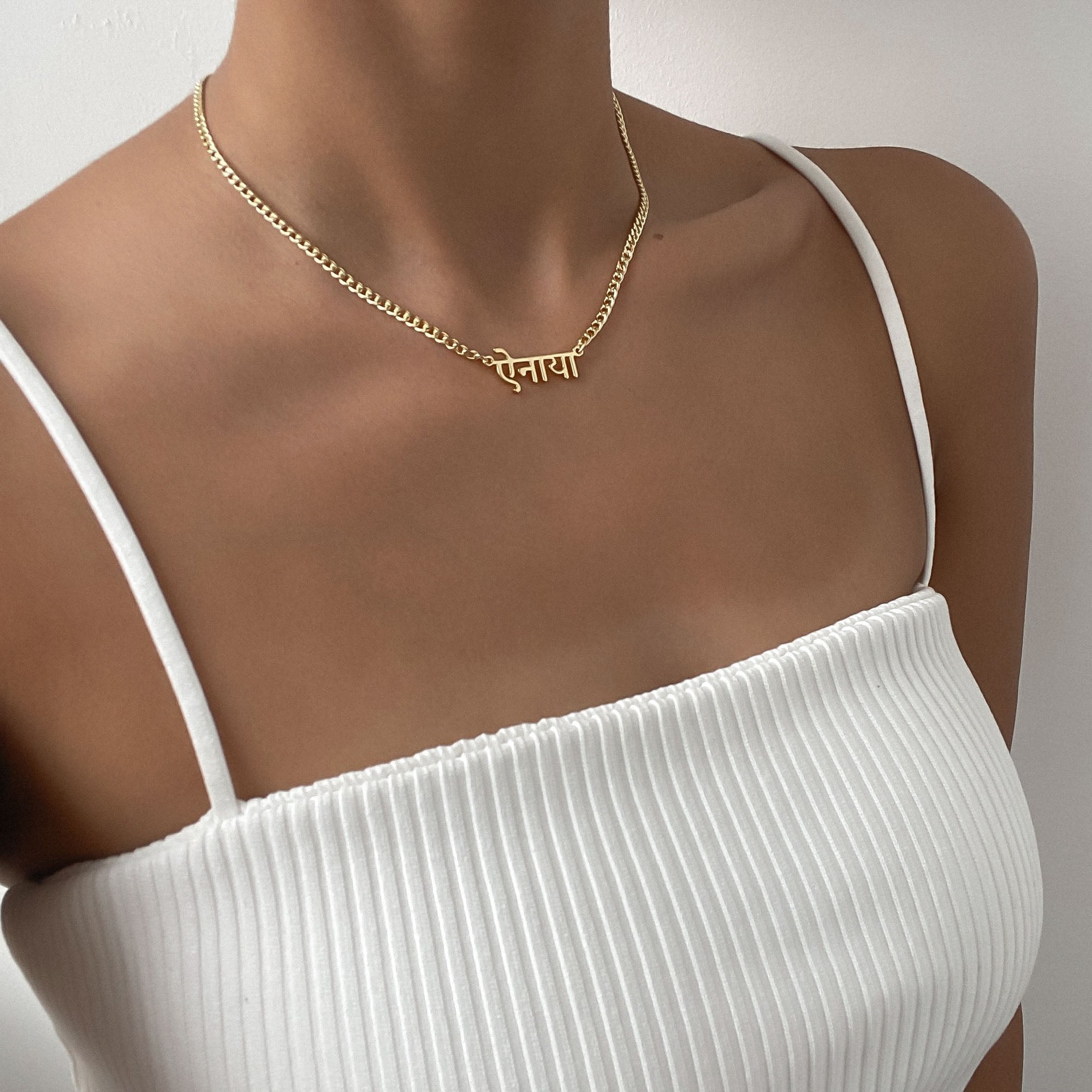 View of a woman’s chest wearing a white strappy top and a gold Hindi name necklace