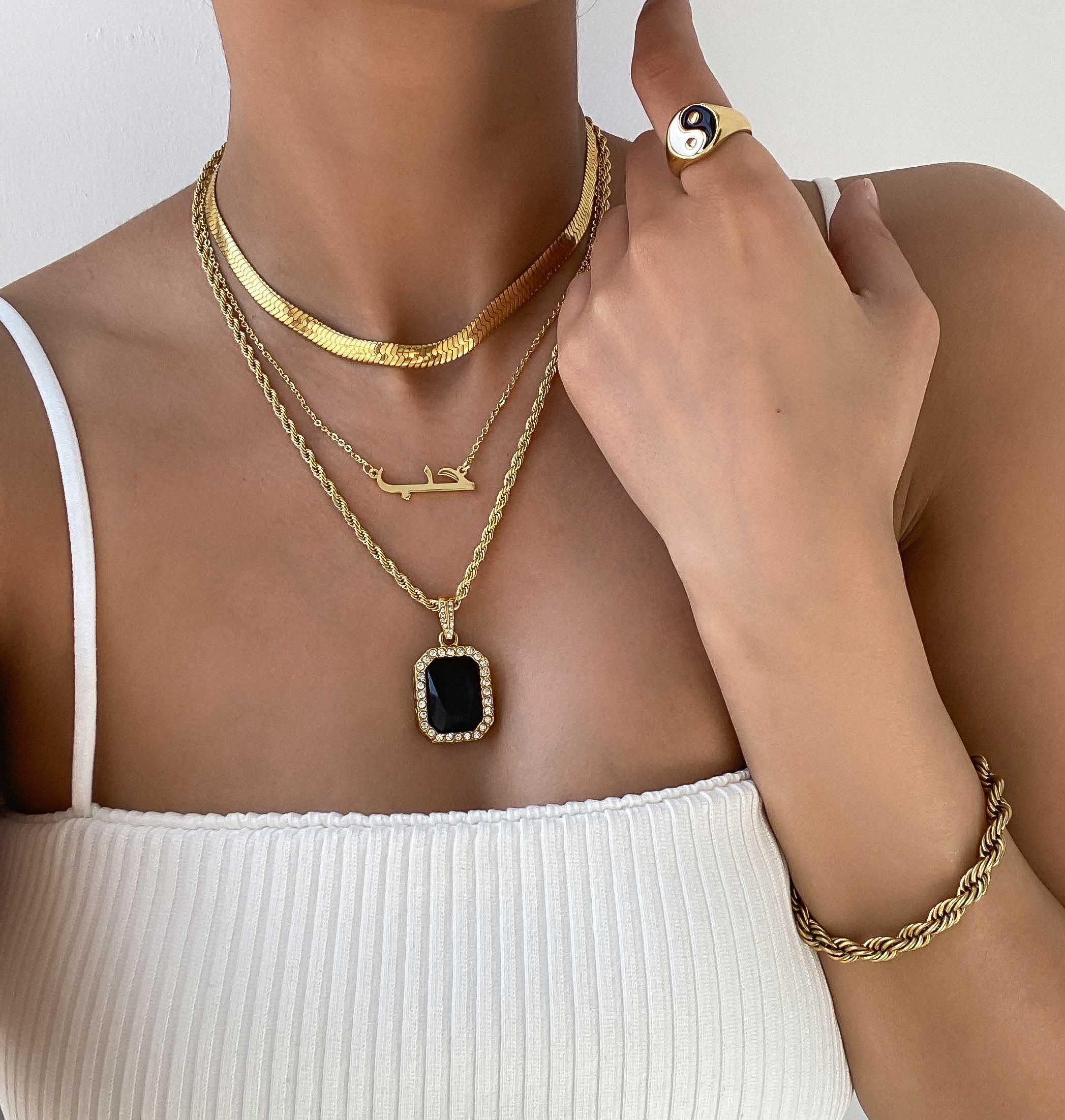 A woman wearing a gold Arabic name necklace layered with an Onyx pendant necklace and Herringbone chain. 